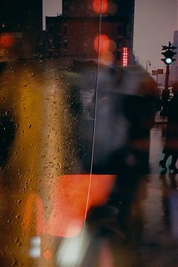 An vertical format photo of a city street scene shot at dusk through a window with raindrops on it. The image is blurry except for the raindrops, and dark in value accented in reds and yellows. Through a reflection a traffic light and a woman in high heels is in focus on the right edge of the frame. An vertical format photo of a city street scene shot at dusk through a window with raindrops on it. The image is blurry except for the raindrops, and dark in value accented in reds and yellows. Through a reflection a traffic light and a woman in high heels is in focus on the right edge of the frame.