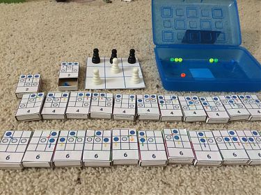 A photo showing a 3x3 chessboard, a set of 24 small boxes with move diagrams labeled on top, and a blue plastic case with some colored beads in it. A photo showing a 3x3 chessboard, a set of 24 small boxes with move diagrams labeled on top, and a blue plastic case with some colored beads in it.