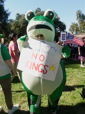 A photo of an individual in an inflatable frog costume at a protest holding a sign reading After 249 years we still believe No Kings.