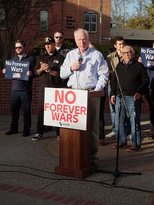 A photo showing an older man with with white hair and a light blue buttonshirt speaking from a podium. The sign under the podium reads NO FOREVER WARS in red letters.