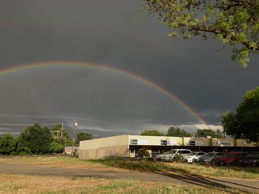A photo of a double rainbow with a low arc above a yellowish one-story commercial building with several for lease signs.