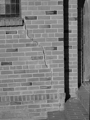 A black and white photograph in portrait orientation of a brick wall with steps in the lower right corner and a window in the upper left corner. There is a crack running diagonally across the wall upper left to lower right.