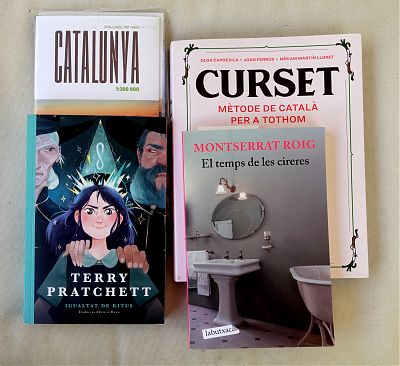 A photo of a map of Catalunya and three books in Catalan, including a novel by Montserrat Roig, a translation of Equal Rites by Terry Pratchett, and a Catalan language textbook.